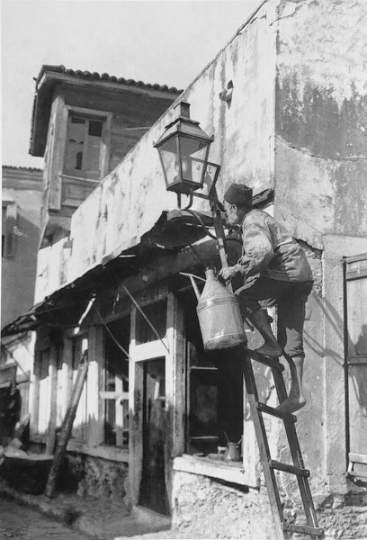 Ο φανοκόρος, εφοδιασμένος με το ειδικό δοχείο του φωτιστικού πετρελαίου, ετοιμάζεται να ανάψει φανάρι.  Το 1862 είχε ολοκληρωθεί το εργοστάσιο φωταερίου στο Γκάζι, ώστε τμήμα των Αθηνών να φωτιστεί με φωταέριο, αλλά στις φτωχογειτονιές επικρατούσε μαύρο σκοτάδι. Απέχουμε ακόμα από την εποχή του Χαρίλαου Τρικούπη.
