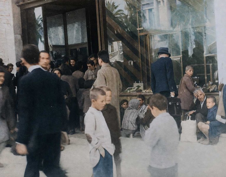 Στην οδό Πανεπιστημίου, μπροστά στο ζαχαροπλαστείο του Τσίτα. Οι μεγάλοι περιμένουν το συσσίτιο, τα παιδιά συμπαραστέκονται και συζητούν.