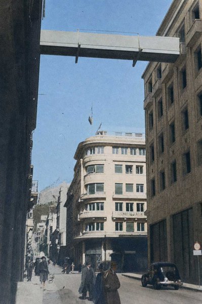 Οδός Αμερικής. Τα κτίρια του Μετοχικού Ταμείου Στρατού και της Αθηναїκής Λέσχης έχουν επιταχθεί από τους Γερμανούς, οι οποίοι για να επικοινωνούν ασφαλώς κατασκεύασαν την εικονιζόμενη γέφυρα. Στο βάθος, στην οδό Πανεπιστημίου, το Commando Piazza, το φρουραρχείο των Ιταλών.
